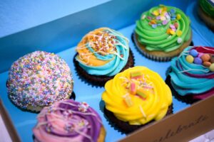 the photo shows a close up of six cupcakes, each decorated with different toppings and various bright colours