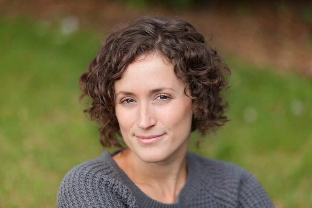 the photo shows a woman with dark curly hair and eyes who is outside and smiling straight at the camera