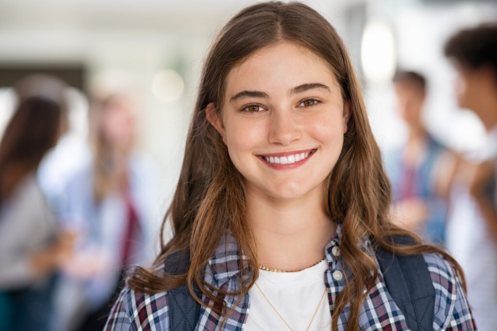 a young girl with dark eyes and long brown hair is wearing a rucksack and smiling straight at the camera